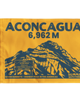 A yellow summit flag featuring a dark blue graphic of the Aconcagua mountain peak, with the text 'ACONCAGUA 6,962 M' at the top and 'SUMMIT SOMETHING' at the bottom right.