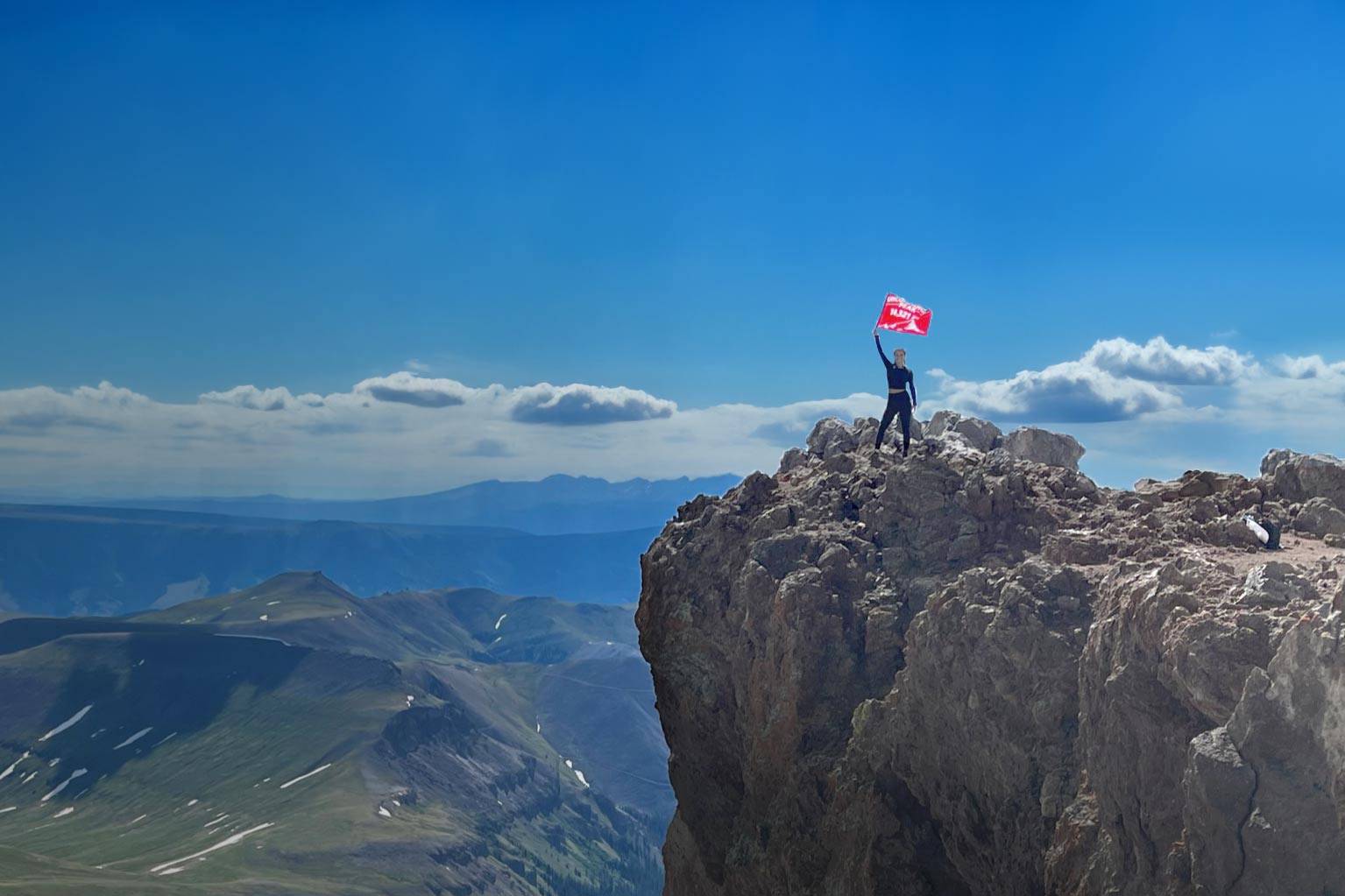 woman on a summit of a mountain holding an orange 5ummit 5omething Summit Flag